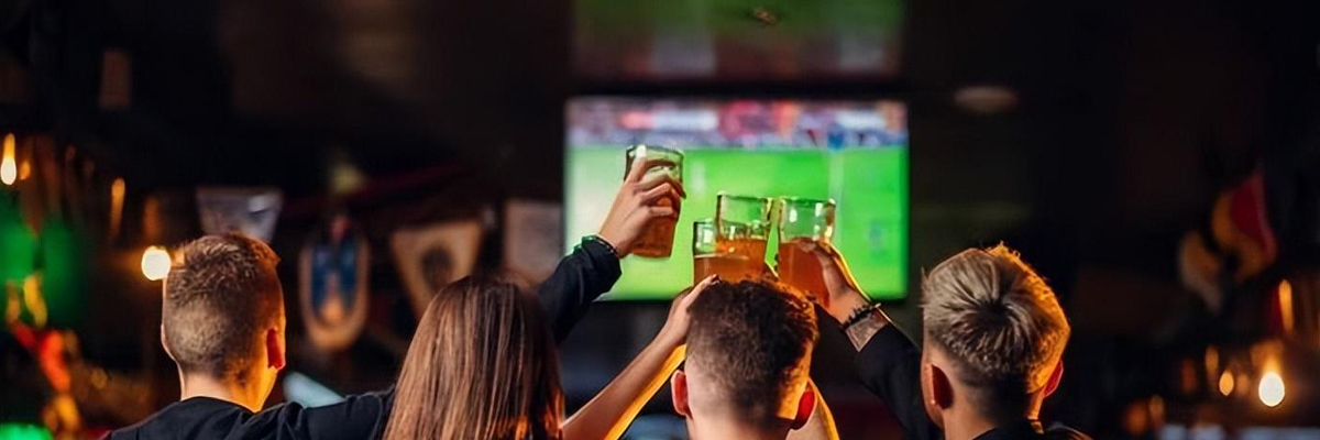 People enjoying drinks and cheering while watching a live World Cup football match on a screen inside a cozy, dimly lit pub or bar